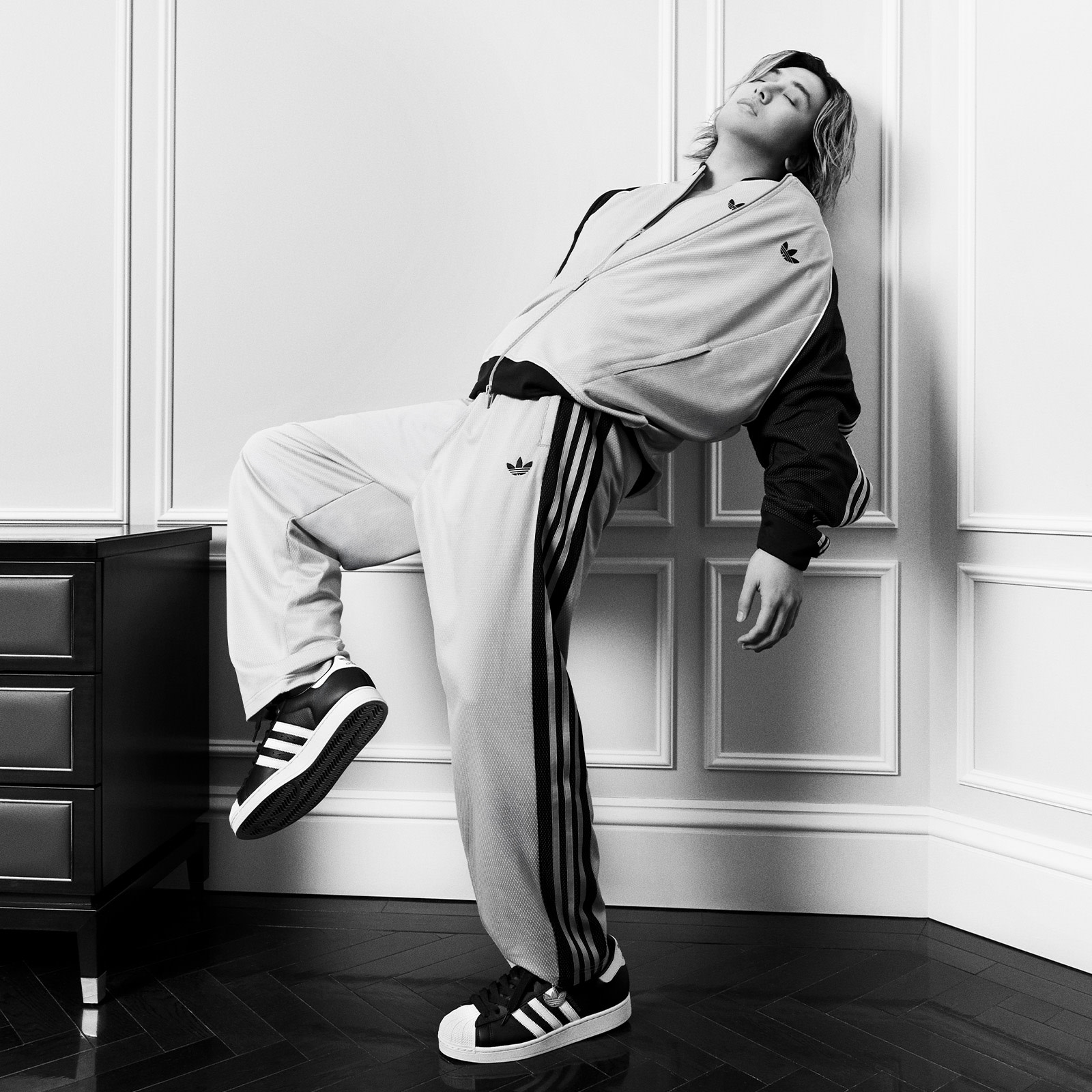 A person in a gray robe and black pants is leaning against a white wall, with a dresser visible in the background.