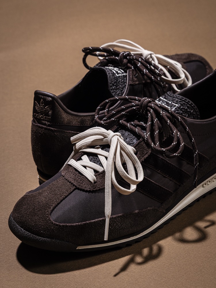 A pair of black and white sneakers with intricate laces and a textured surface, set against a plain brown background.