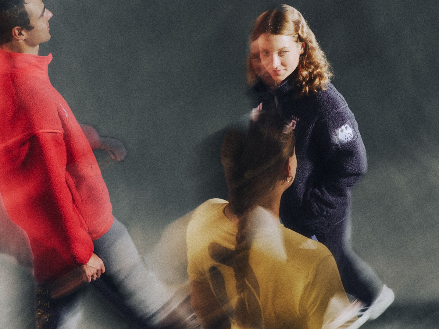 Zwei Personen, eine mit rotem und die andere mit dunklem Kleidungsstück, stehen in einem dynamischen Zusammenspiel vor einem dunklen Hintergrund.