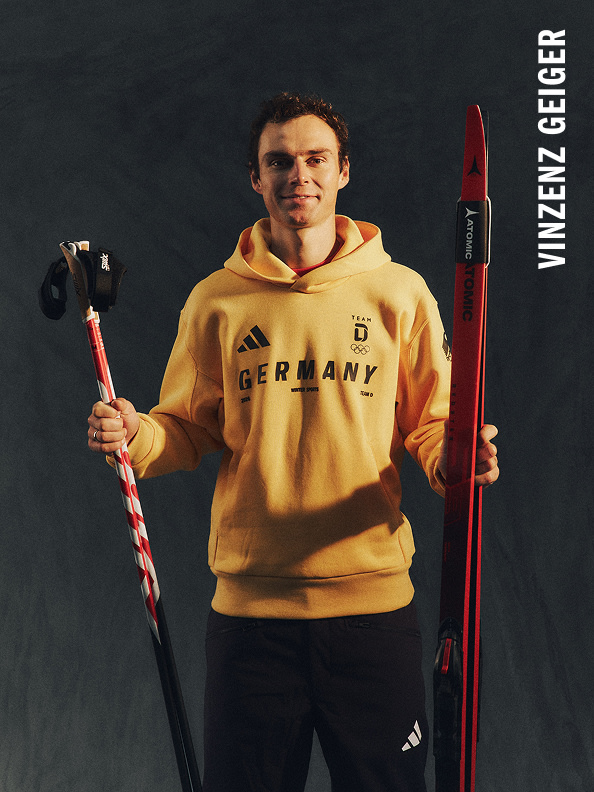 Ein junger Mann in einem gelben Hoodie mit „Germany“-Print, der einen Skistock und einen roten Skistock vor einem dunklen Hintergrund hält.