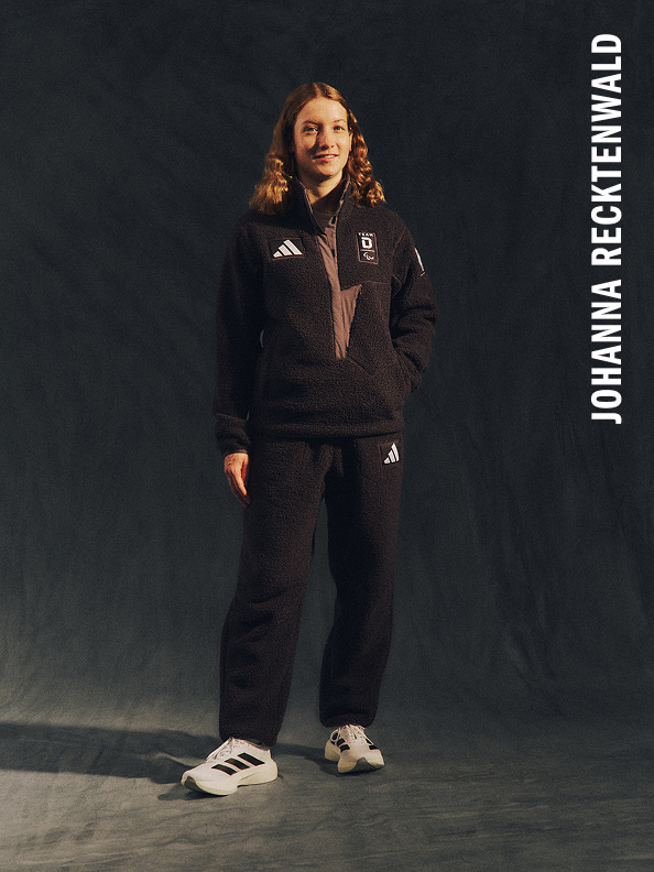 Eine junge Frau in einem adidas Trainingsanzug steht in einer dunklen, schattigen Umgebung. Ihre Ausdruckskraft und Haltung vermittelt ein Gefühl von Entschlossenheit.