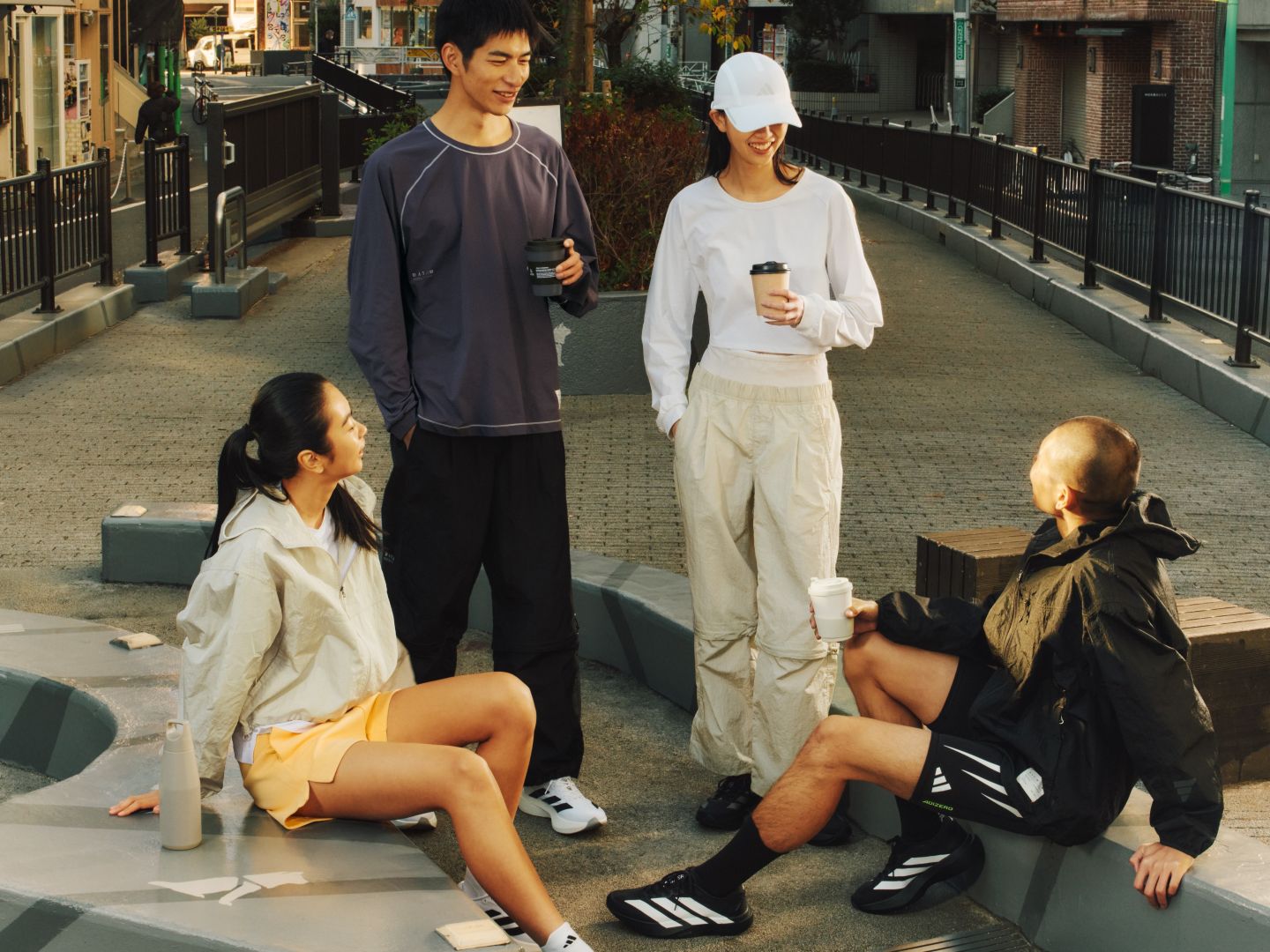The image depicts a group of people, some sitting on the ground and others standing, in an urban setting with buildings and a walkway visible in the background.