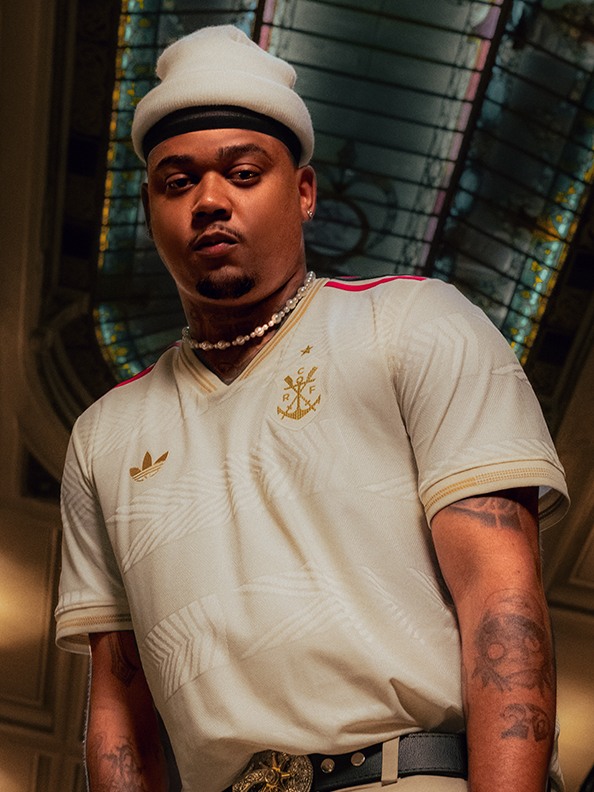 A man wearing a white hat and shirt with a tattoo on his arm stands in front of a colorful, patterned background.