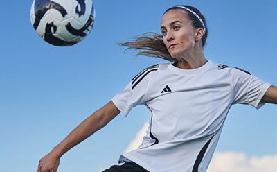 A female soccer player in a white jersey is seen kicking a soccer ball against a clear blue sky.