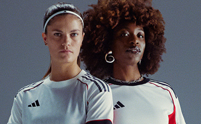 Two individuals, one with short light-colored hair and the other with curly dark hair, are wearing white athletic jerseys against a plain gray background.