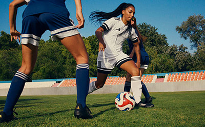 Two female soccer players in uniform are competing for the ball on a grassy field, with a blue sky and trees visible in the background.