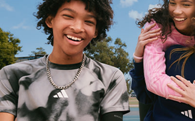 Two young people, a boy and a girl, are smiling and laughing together outdoors in a park-like setting with trees and a blue sky in the background.