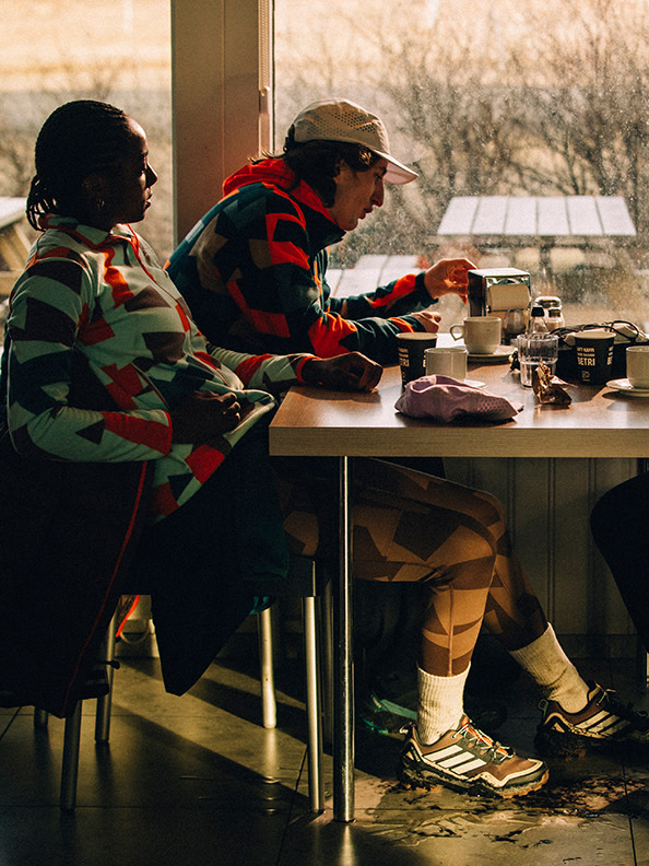 Two individuals, one wearing a camouflage jacket and the other a red and black patterned jacket, are seated at a table in what appears to be an outdoor setting, with a solar panel visible in the background.
