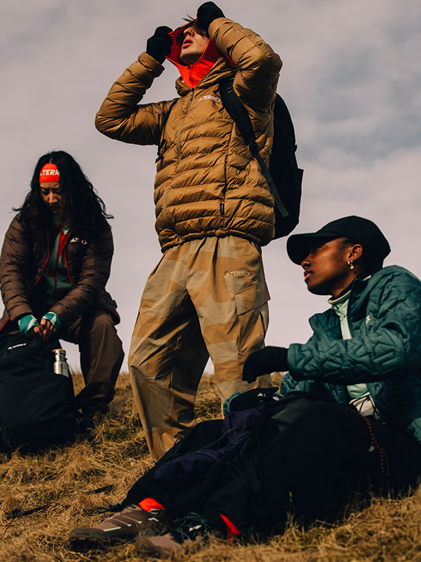 Three individuals dressed in warm winter attire are gathered in a grassy field against a cloudy sky backdrop.