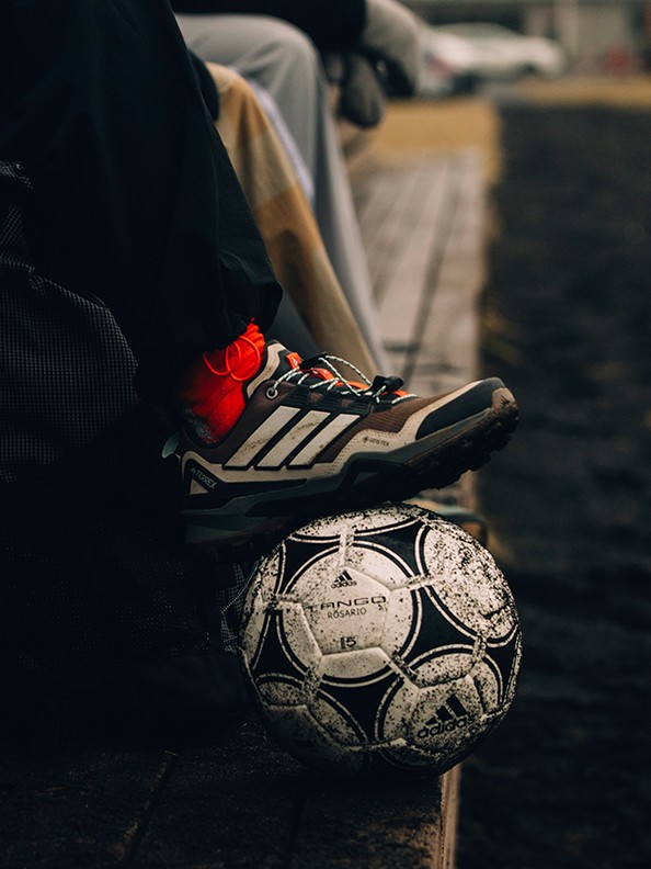 A worn soccer ball rests on the ground, with a pair of cleats visible in the foreground, suggesting a scene of a soccer player or coach preparing for a game or practice.