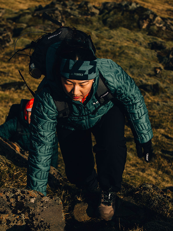 A person in warm outdoor gear crouches on a rocky, rugged terrain with a mountainous backdrop.