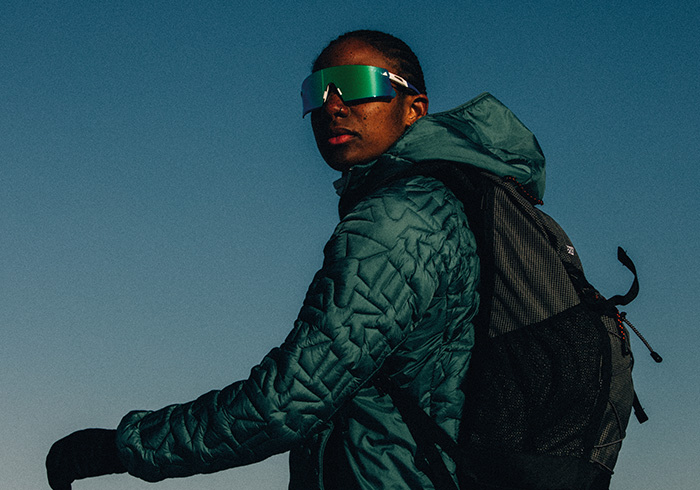 A person wearing a green quilted jacket and dark sunglasses stands against a clear blue sky.