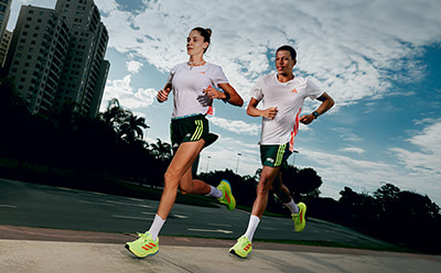 Two runners in athletic gear are jogging on a paved path surrounded by tall buildings and palm trees, with a bright blue sky overhead.