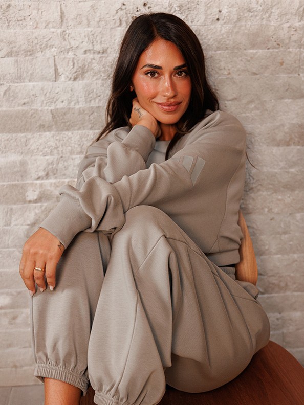A young woman with dark hair sits comfortably on the floor, wearing a cozy beige outfit, against a plain white brick wall background.