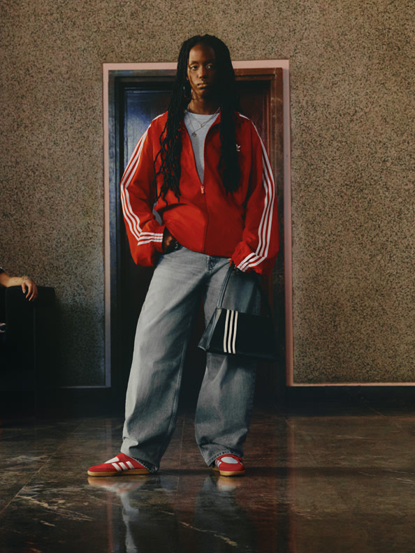 A person wearing a red jacket and gray sweatpants stands in a dimly lit room with a plain wall in the background.