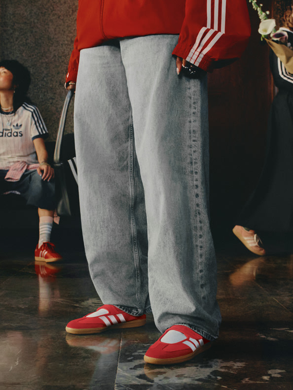 The image shows a person wearing red Adidas clothing and gray sweatpants, standing on a wooden floor with other people in the background.