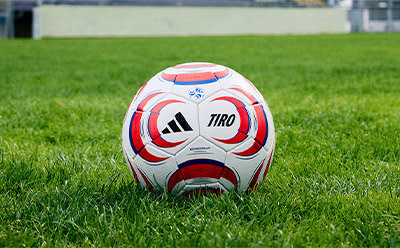A white and red soccer ball sits on a lush green grass field, with a plain white building visible in the background.