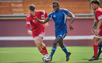 The image depicts two soccer players, one in a red uniform and the other in a blue uniform, engaged in a competitive match on a grassy field with a stadium-like structure visible in the background.