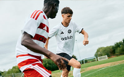Two soccer players, one in a white jersey and the other in a red and white striped jersey, are engaged in a match on a grassy field with a goal post visible in the background.