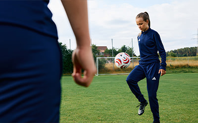 A person in a blue sports uniform is kicking a soccer ball on a grassy field, with a rural landscape visible in the background.