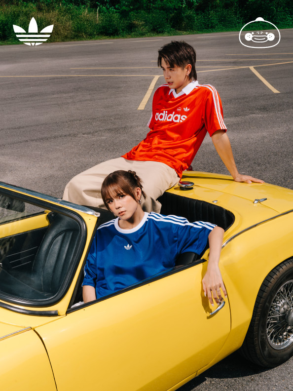 Two individuals, a male and a female, are seated in a yellow sports car against a backdrop of a grassy area and trees.