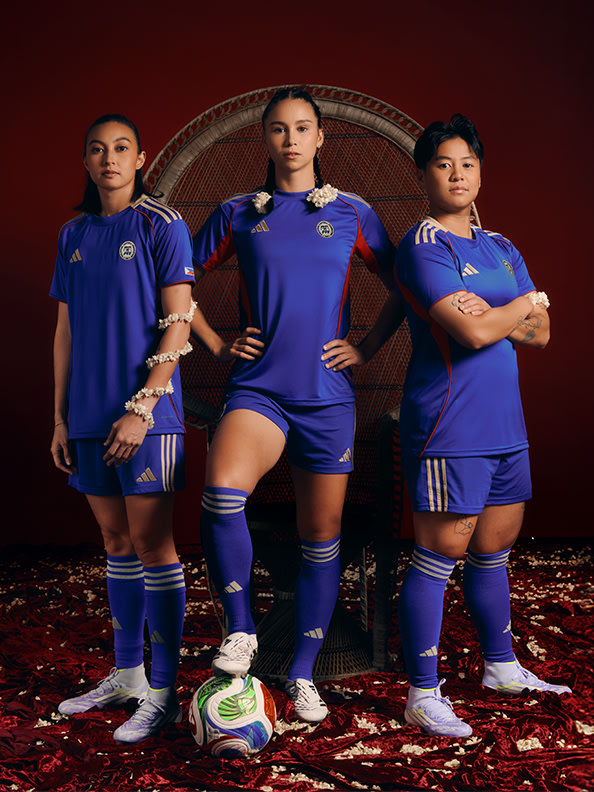 Three female soccer players in blue uniforms standing in front of a woven background, with a soccer ball at their feet.