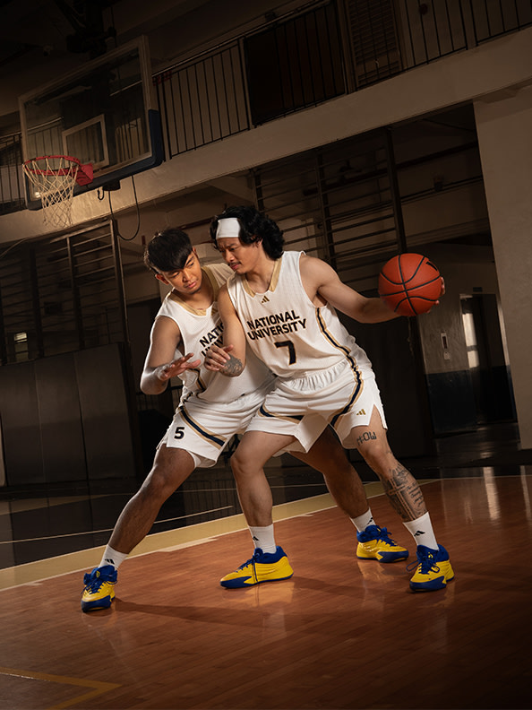Two basketball players in jerseys are facing each other on a basketball court, with a basketball hoop visible in the background.