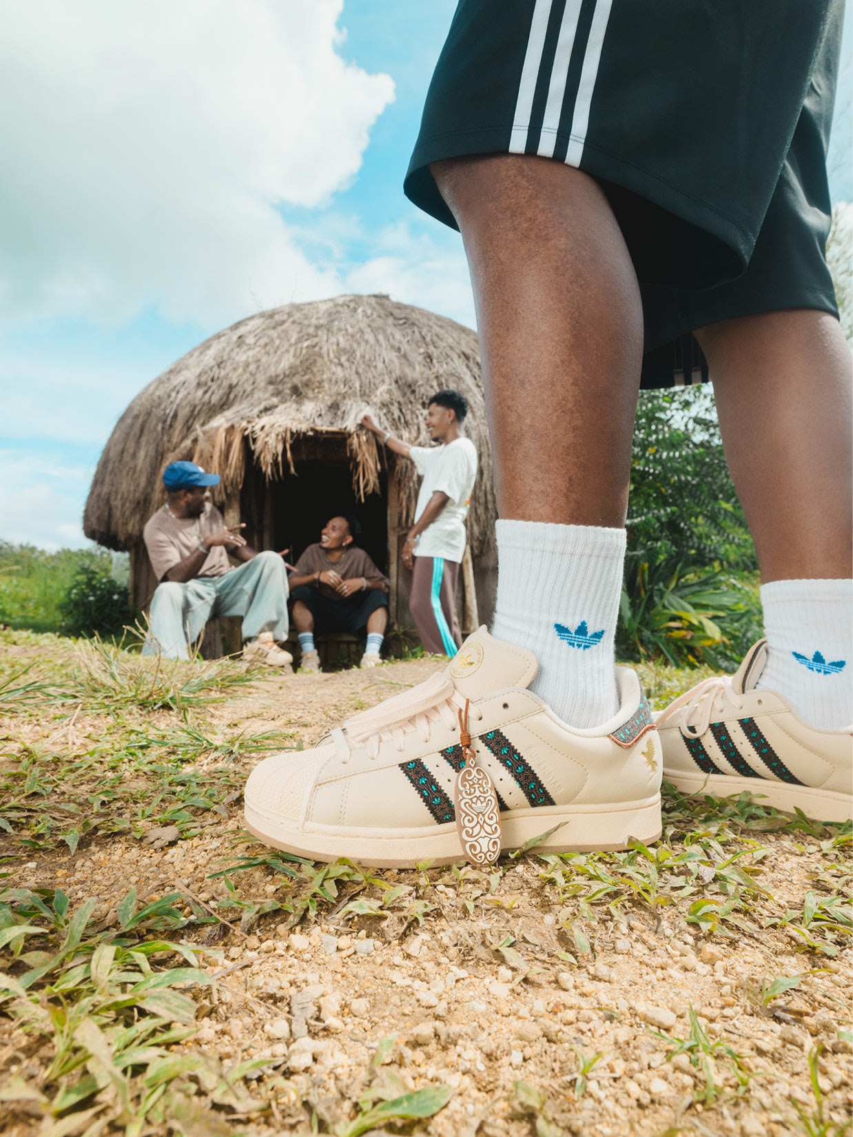 The image shows a person wearing Adidas sneakers standing in a grassy field with a traditional hut-like structure in the background, surrounded by other people.