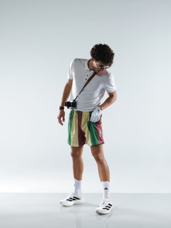 A person with curly hair wearing a white t-shirt, colorful shorts, and white sneakers stands against a plain light background.