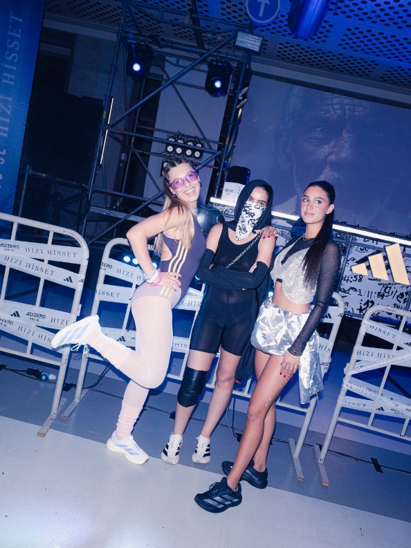 Three young women posing together in front of a stage with blue lighting and equipment in the background.