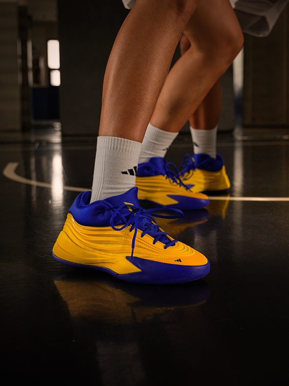 The image shows a pair of bright yellow and blue sneakers with laces, worn by someone standing on a dark surface.