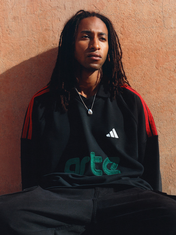 A person with dreadlocks wearing a black and red Adidas jacket stands in front of an orange-tinted wall.