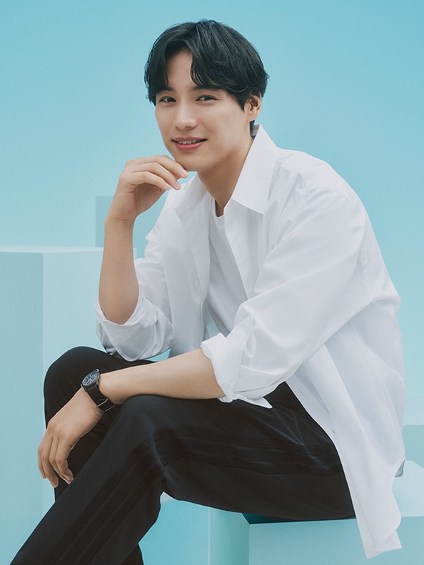 A young man in a white shirt and black pants sits on a blue background, smiling warmly at the camera.