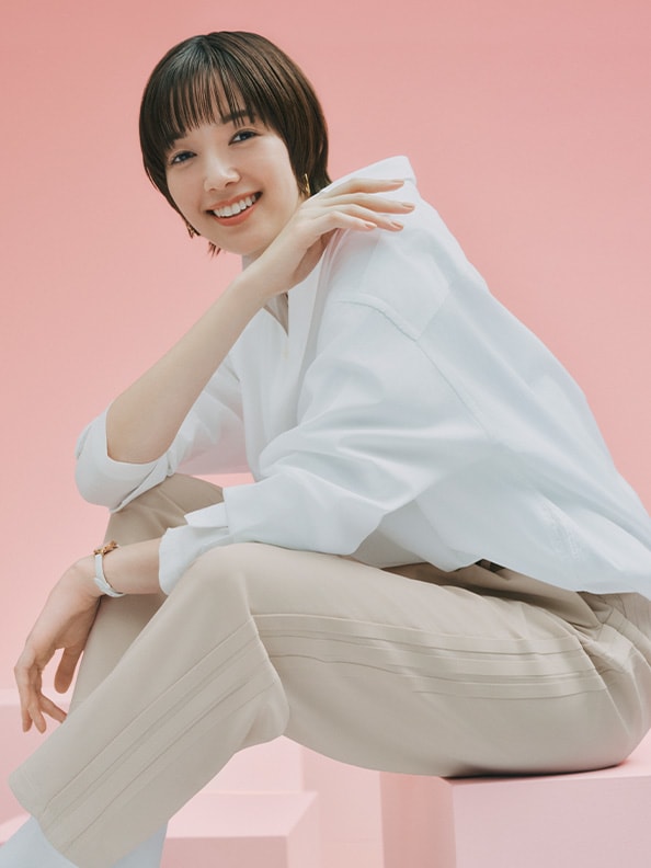 A smiling young woman in a white top and beige pants sits on a pink background.