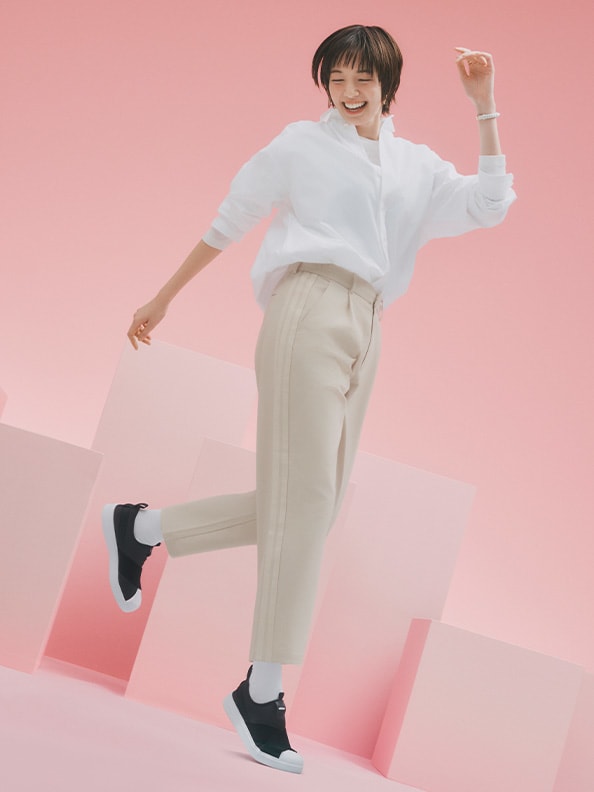 A person wearing a white sweater and beige pants is jumping excitedly against a pink background.
