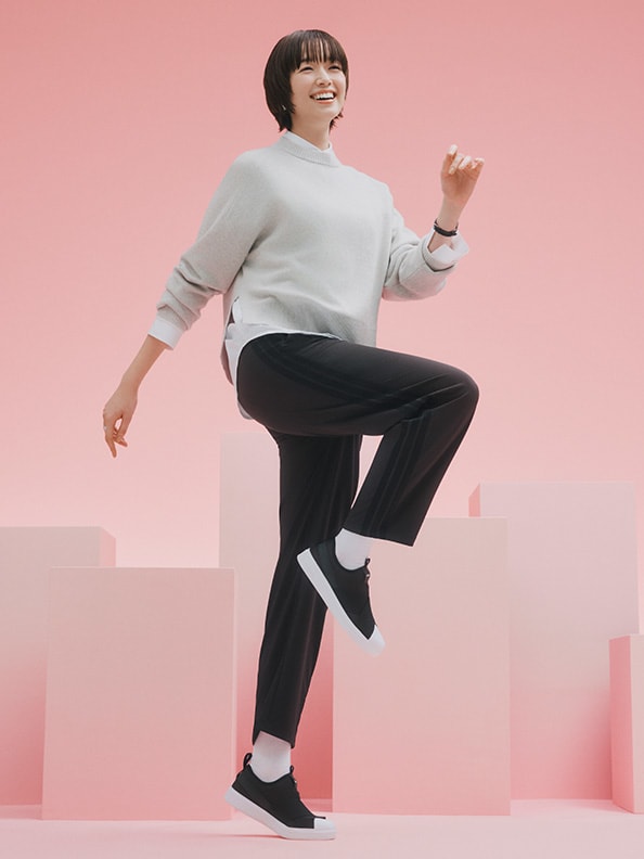 A young woman in a gray sweater and black pants is standing in a pink-toned environment, appearing energetic and joyful.