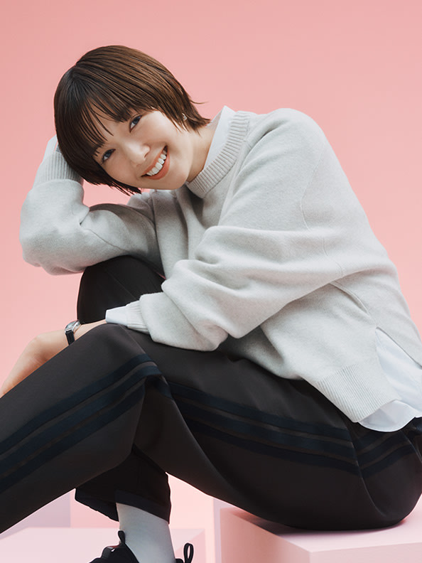 A young woman with short, dark hair is sitting on the floor, wearing a white sweater and black pants, against a pink background.