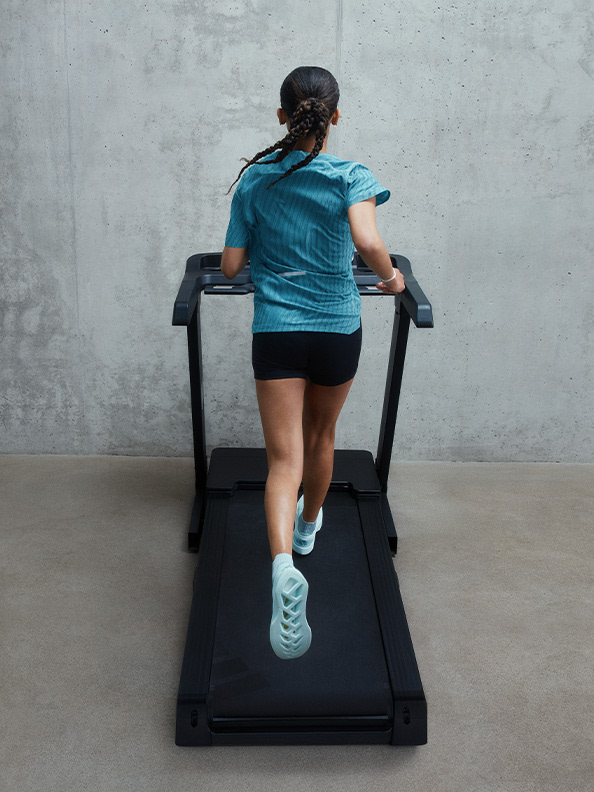 A person in a blue shirt and black shorts is walking on a treadmill in a room with a concrete wall in the background.