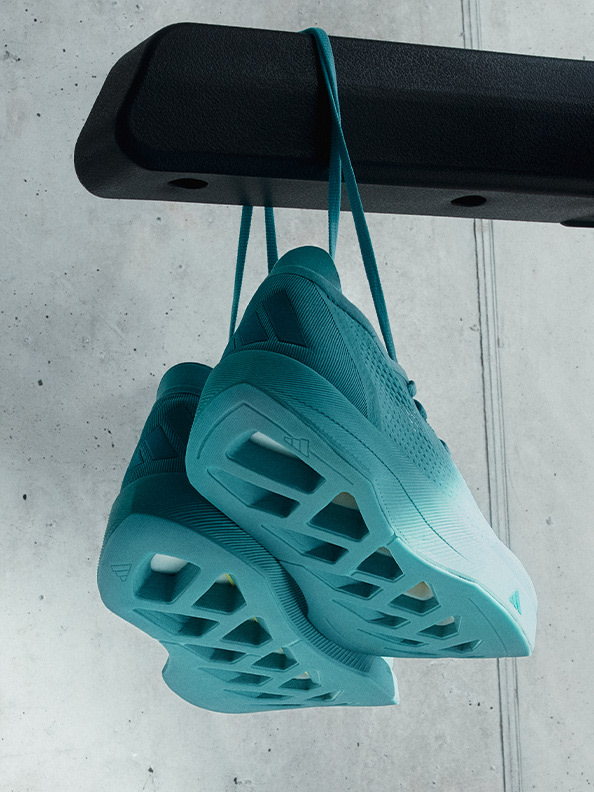 A pair of teal-colored, futuristic-looking sneakers are suspended from a black metal rack against a plain concrete wall.