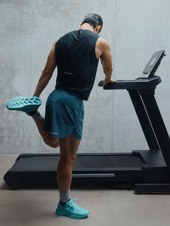 A muscular man in athletic attire stands on a treadmill, holding a green exercise mat, with a plain concrete wall in the background.