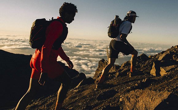 Two people in hiking gear stand on a rocky mountain ridge, with a vast expanse of clouds and sky visible in the background.
