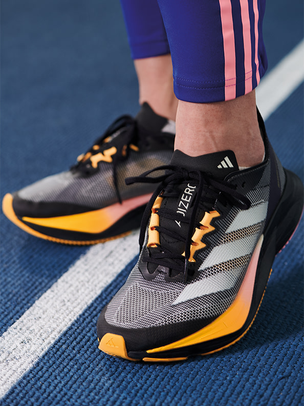 L'image montre une paire de chaussures de sport grises et orange avec le logo adidas sur le côté, reposant sur une surface bleue et blanche.