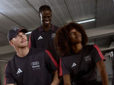The image shows three individuals wearing black sports jerseys with the Audi logo, standing in what appears to be a sports facility or training center.