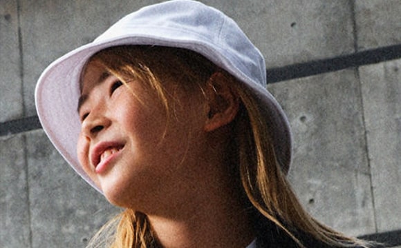 A young person with long, light-colored hair wearing a white hooded sweatshirt, their face partially obscured, standing in front of a concrete wall.