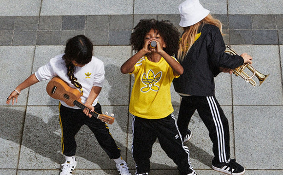 Three individuals, two women and one man, are standing in an urban setting with a tiled wall in the background. The woman in the center is wearing a yellow shirt and appears to be singing or performing, while the other two individuals are holding musical instruments.