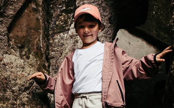 A young person wearing a pink jacket and hat stands in front of a rocky, wooded area, smiling at the camera.