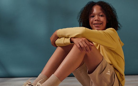 A young woman with curly dark hair sits on the floor, wearing a yellow top and beige pants, against a teal background.