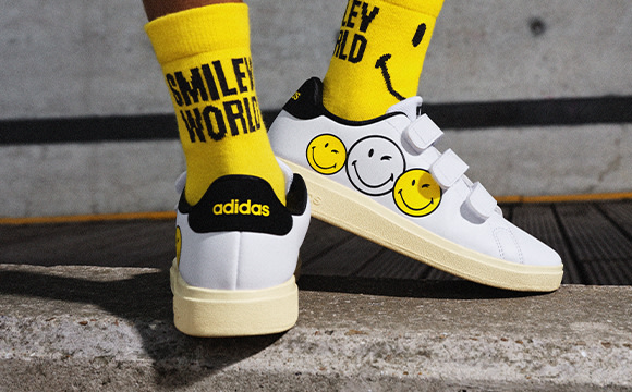 The image shows a pair of white Adidas sneakers with yellow socks featuring smiley face designs, standing on a concrete surface.