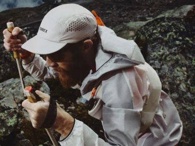 A person wearing a white cap and a gray jacket is standing in a rocky, mountainous landscape.