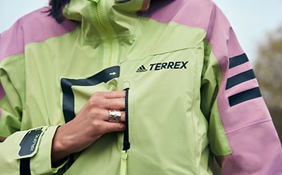 Das Bild zeigt eine Person, die eine hellgrün-pinke adidas TERREX Jacke trägt, wobei ihre Hand im Vordergrund zu sehen ist.